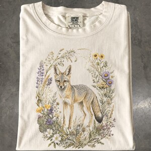 Peut inclure: Un t-shirt à col rond couleur crème avec une impression graphique représentant un renard au centre d'une couronne de fleurs sauvages et d'herbes. L'étiquette intérieure du col affiche le texte Comfort Colors et la taille L-L-L.