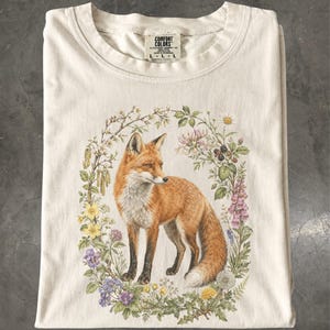 Puede incluir: Una camiseta de color crema con un estampado gráfico de un zorro rojo de pie dentro de una corona ovalada de flores silvestres coloridas, bayas y follaje verde. La etiqueta interior del cuello muestra el texto Comfort Colors y la talla L-L-L.