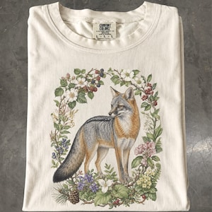 Peut inclure: Un t-shirt couleur crème avec une étiquette Comfort Colors au col. L'illustration représente un renard gris et orange debout au centre d'une couronne circulaire détaillée composée de fleurs sauvages, de feuilles, de baies et d'une pomme de pin.