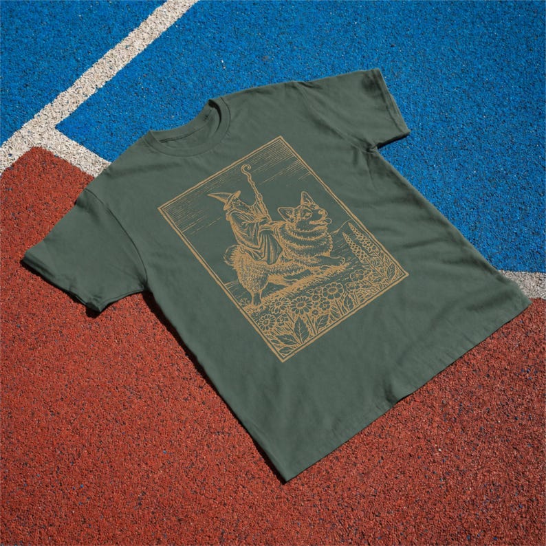 May include: An olive green short-sleeved t-shirt featuring a tan line-art graphic of a robed figure wearing a pointed hat and holding a staff while riding a large dog, all framed within a rectangular border surrounded by floral elements.