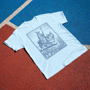 May include: A light blue short-sleeve t-shirt featuring a dark blue graphic print of a wizard riding a corgi. The illustration is framed in a rectangular border and depicts the wizard holding a staff while surrounded by flowers and foliage.