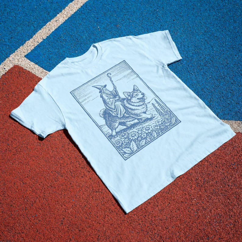 May include: A light blue short-sleeve t-shirt featuring a dark blue graphic print of a wizard riding a corgi. The illustration is framed in a rectangular border and depicts the wizard holding a staff while surrounded by flowers and foliage.