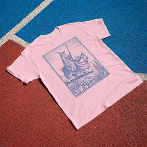 May include: A light pink short-sleeved t-shirt featuring a dark blue rectangular graphic print. The illustration depicts a wizard wearing a pointed hat and robe riding on the back of a corgi dog, surrounded by flowers and tall plants.