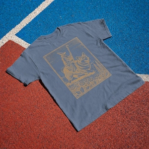 May include: A slate blue short-sleeved t-shirt featuring a gold-colored line art graphic of a wizard figure riding a corgi dog. The illustration is framed within a rectangular border and includes detailed floral elements at the base of the design.