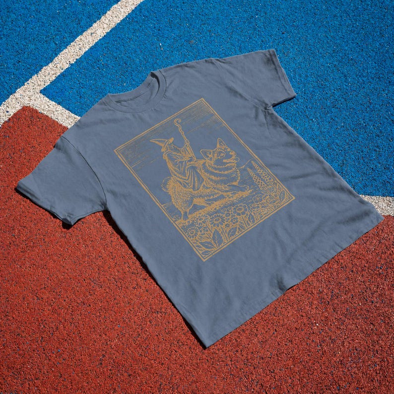May include: A slate blue short-sleeved t-shirt featuring a gold-colored line art graphic of a wizard figure riding a corgi dog. The illustration is framed within a rectangular border and includes detailed floral elements at the base of the design.