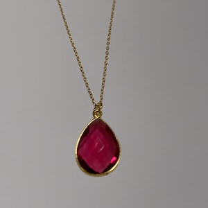 May include: A gold-colored necklace with a teardrop-shaped, faceted, ruby-red pendant. The pendant is framed in gold and hangs from a delicate gold chain. The necklace is displayed against a plain, light background.