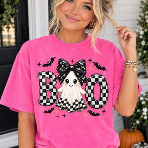 May include: A bright pink short-sleeve t-shirt featuring a graphic print of the word BOO with a ghost wearing a large black sequined bow in the centre. The letters B and O are filled with a black and white chequered pattern, surrounded by small black bats.