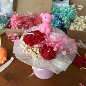 Könnte beinhalten: Ein kleines Kerzenarrangement mit einem rosa Teddybären, roten und rosa rosenförmigen Kerzen und Schleierkraut. Das Arrangement ist in weißes Papier gewickelt und mit einem weißen Band gebunden, präsentiert in einem rosa Zylinder.
