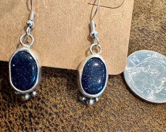 May include: Silver-toned dangle earrings with oval, dark blue, speckled stones. The stones are set in a silver bezel with a small decorative cluster at the bottom. The earrings hang from a simple silver hook. The background includes a brown surface and a cardboard earring display card.
