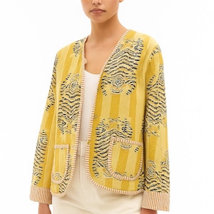May include: Mustard yellow jacket featuring a tiger stripe design. The jacket has a V-neck, two front pockets, and a striped trim. The tiger stripes are black and the background is yellow. The jacket is worn over a white top and paired with white trousers.