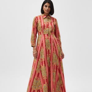 Op de afbeelding: Een lange, knoopsluiting jurk met een rood-roze streepmotief en tijgerprint accenten. De jurk heeft lange mouwen en een kraag. Het model draagt gouden oorbellen en sandalen.