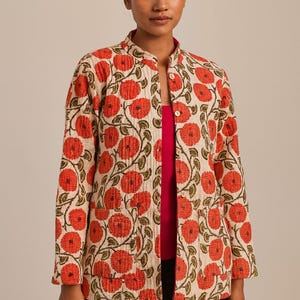 May include: A cream-colored jacket with a vibrant floral pattern of red flowers and green leaves. The jacket has a mandarin collar, long sleeves, and two front pockets. The model is wearing a pink top and black pants.