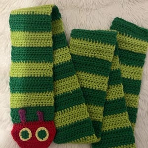 May include: A handmade crochet scarf with green and light green stripes. The scarf features a red caterpillar head with purple antennae and yellow eyes. The scarf is made of soft yarn.