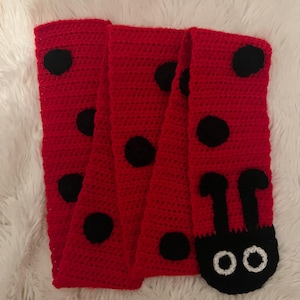 May include: A red crocheted scarf with black polka dots and a ladybug head design at the end. The scarf is made of a soft, textured yarn and is displayed on a white, fluffy surface. The ladybug head features white eyes and black antennae.