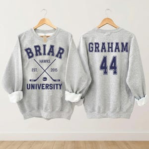 Op de afbeelding: Twee grijze sweatshirts op houten hangers. Het sweatshirt aan de voorkant heeft "Briar University" in marineblauwe letters met gekruiste hockeysticks. Het sweatshirt aan de achterkant heeft "Graham 44" in marineblauwe letters.