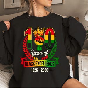 Könnte beinhalten: Schwarzes Sweatshirt mit einer Grafik, die 100 Jahre Black Excellence feiert. Das Design zeigt die Zahl 100 in Rot, Gelb und Grün, eine Faust, eine Krone und den Text "Years of Black Excellence 1926-2026."