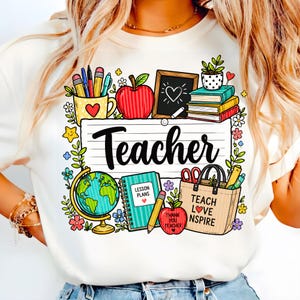 May include: White t-shirt with a colorful graphic design featuring school-related elements. The design includes an apple, books, a globe, and the word "Teacher" in large letters. Additional text includes "Lesson Plans" and "Teach Love Inspire".