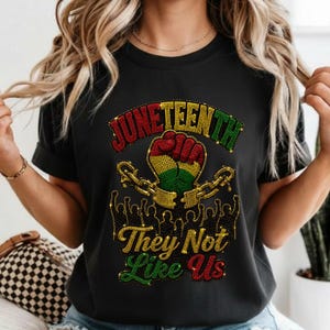 May include: Black t-shirt with a Juneteenth graphic. The design features the word "Juneteenth" in red and green, a clenched fist, and the text "They Not Like Us". The graphic includes gold chains and silhouettes.