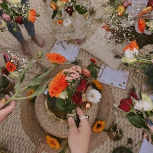 Op de afbeelding: Een verzameling kleurrijke boeketten met oranje gerbera's, rode rozen en witte bloemen. De boeketten zijn gerangschikt op een geweven mat, sommige worden door mensen vastgehouden. De scène wordt verlicht door natuurlijk licht, wat een warme sfeer creëert.