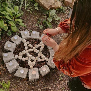 May include: A circular arrangement of gray stone blocks and white pebbles, with small candles in glass holders. The stones are set in a circle, with a star-shaped pattern of pebbles in the center. The scene is set outdoors, surrounded by greenery.