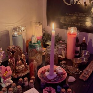 May include: A collection of items including a tall pink candle in a pink dish, a gold-coloured Buddha statue, several glass jars filled with small objects, a light blue pillar candle, a pink pillar candle, and various small crystals and trinkets.