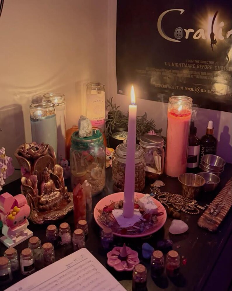May include: A collection of items including a tall pink candle in a pink dish, a gold-coloured Buddha statue, several glass jars filled with small objects, a light blue pillar candle, a pink pillar candle, and various small crystals and trinkets.
