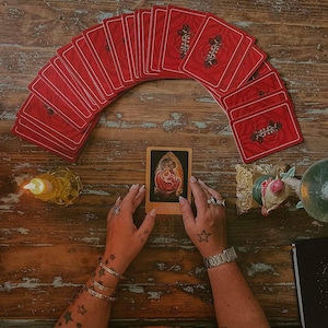 May include: A tarot card reading setup featuring a deck of red-backed cards arranged in an arc. A single card is displayed, depicting a figure within an ornate frame. The scene includes a lit candle and decorative items on a wooden surface.