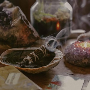 May include: A smoldering sage bundle rests in an abalone shell, releasing wisps of smoke. The scene includes crystals, a small terrarium, and tarot cards, suggesting a spiritual or wellness theme. The items are arranged on a wooden surface.