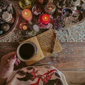 以下が含まれることがあります： コーヒーカップ、手書きのテキストが書かれた開いたジャーナル、キャンドル、装飾品を備えたボヘミアンな静物画。ジャーナルのテキストには「私は平和で、私は強い」と書かれています。手がマグカップを持っています。画像は暖かく、魅力的な美学を持っています。