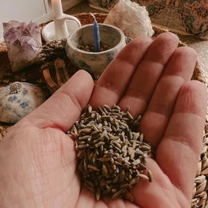 Könnte beinhalten: Nahaufnahme einer Hand, die getrocknete Lavendelblüten hält, grün und braun. Im Hintergrund Kristalle, eine Kerze und eine kleine Keramikschale mit einer blauen Kerze.