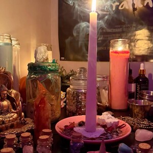May include: A tall, light pink taper candle burns on a small pink plate, surrounded by various glass jars, a gold-coloured Buddha figurine, small crystals, and a pink flower-shaped incense holder with a cone incense on a dark surface.