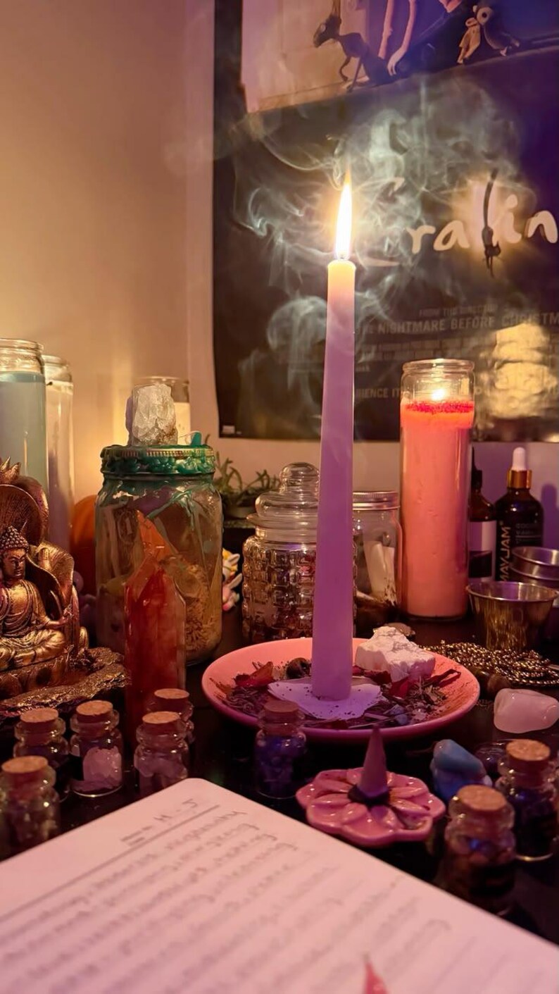 May include: A tall, light pink taper candle burns on a small pink plate, surrounded by various glass jars, a gold-coloured Buddha figurine, small crystals, and a pink flower-shaped incense holder with a cone incense on a dark surface.