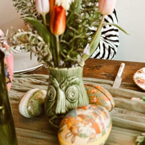 May include: A green tiki-style vase holds a bouquet of tulips in shades of pink, orange, and white. Decorative Easter eggs with floral and animal designs are arranged on a green cloth. A butter knife and a plate are also visible.
