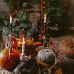 May include: A rustic table setting with a witchy theme. Features include lit candles, pumpkins, a cauldron, a crystal ball, and jars of herbs. The scene is decorated with plants and a decorative antler.