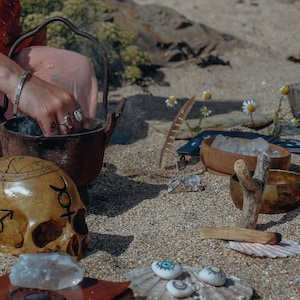 以下が含まれることがあります： 砂浜での神秘的なシーン。湯気を出す鋳鉄製の鍋、マークの付いた頭蓋骨、クリスタル、木製のボウル、ペイントされた石が鍋の周りに配置されています。羽とデイジーが自然な美しさを添えています。