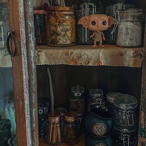 May include: A wooden cabinet filled with glass jars containing herbs and spices. A Funko Pop figure of Dobby from Harry Potter is on a shelf. The jars have metal lids and handwritten labels. The cabinet has a vintage, rustic appearance, with a floral patterned shelf liner.
