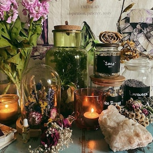 May include: A still life arrangement featuring glass jars, candles, and dried flowers. Jars contain sage, lavender, and other botanicals. A glass cloche displays dried flowers. The scene is illuminated by candlelight, creating a warm, inviting atmosphere.