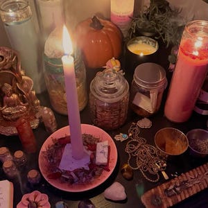 May include: A collection of spiritual items including a lit taper candle in a pink dish, several glass jar candles, a small orange pumpkin, various crystals, a gold-toned hamsa necklace, small glass vials, and a wooden incense burner on a dark surface.