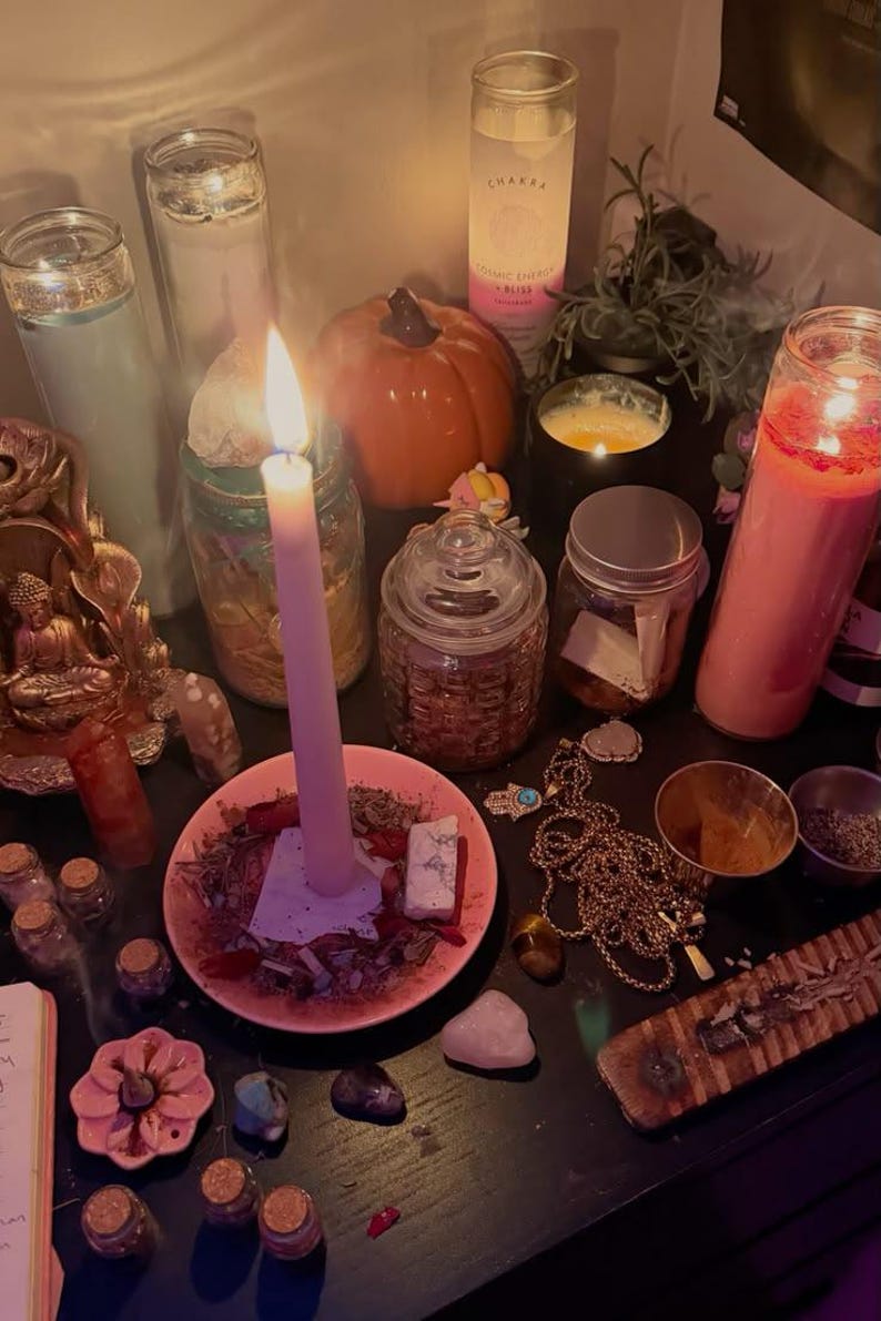 May include: A collection of spiritual items including a lit taper candle in a pink dish, several glass jar candles, a small orange pumpkin, various crystals, a gold-toned hamsa necklace, small glass vials, and a wooden incense burner on a dark surface.