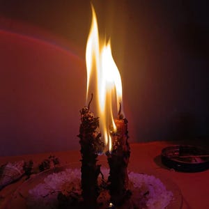 May include: Two dark, textured candles are burning with bright yellow and orange flames. The candles are positioned upright on a clear glass plate covered in a layer of white granular salt, with a small pool of melted wax visible at the base.