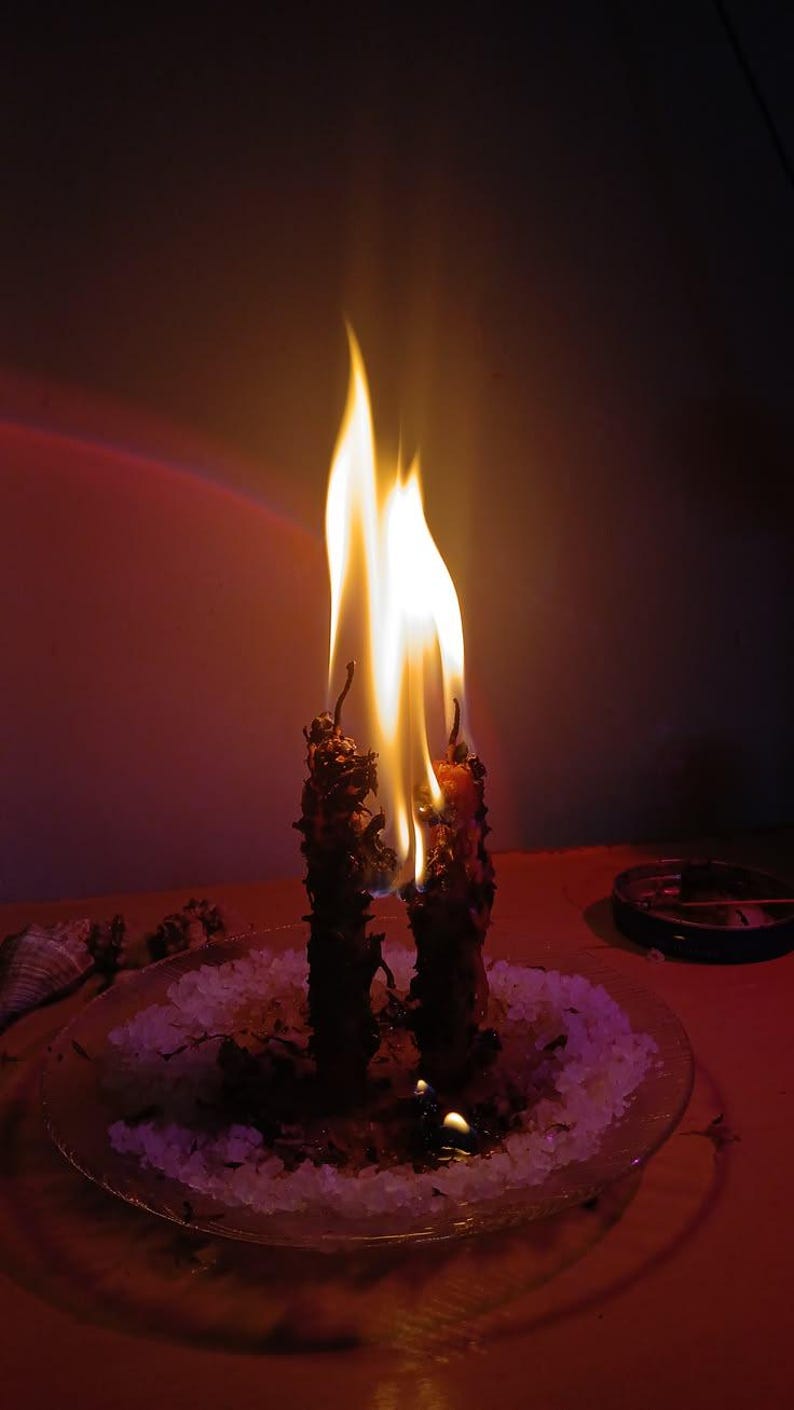 May include: Two dark, textured candles are burning with bright yellow and orange flames. The candles are positioned upright on a clear glass plate covered in a layer of white granular salt, with a small pool of melted wax visible at the base.