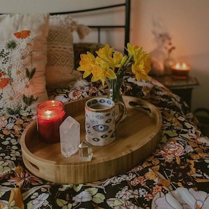 May include: A wooden tray holds a lit red candle, a clear quartz crystal, and a mug with an eye pattern. A vase of yellow daffodils sits next to the mug. The tray rests on a floral patterned bedspread, creating a cozy, bohemian aesthetic.