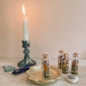 Può includere: Una candela bianca accesa in un candelabro verde acqua, con piccole bottiglie di vetro piene di erbe e cristalli. Una ciotola decorativa color oro contiene una delle bottiglie. Sono presenti anche un cristallo blu e una pietra dipinta con un disegno a occhio.