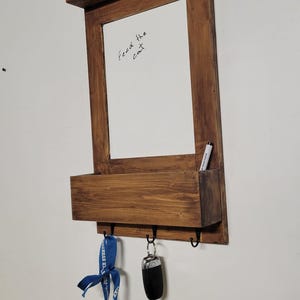 Puede incluir: Un organizador de pared de madera con una pizarra, un estante y ganchos para llaves. La pizarra tiene el texto escrito a mano "Feed the cat". Un cordón azul y una llave de coche cuelgan de los ganchos. Un marcador está en un pequeño compartimento.