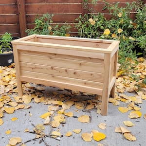 May include: A rectangular wooden planter box with a natural finish. The planter has a slatted design on the sides and is elevated on four legs. The background features plants in black pots and fallen yellow leaves.
