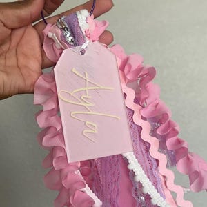 May include: A pink and purple accessory with the name "Ayla" on a tag. The accessory features ribbons, lace, and ruffled fabric in shades of pink and purple. A silver-coloured ring is at the top.