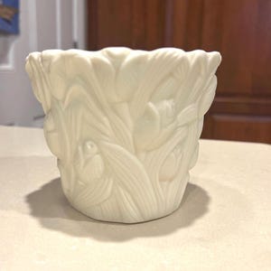May include: Off-white ceramic planter with a detailed relief of tulips and leaves. The planter has a scalloped rim and a rounded base. The design is consistent around the entire planter, creating a cohesive floral theme.