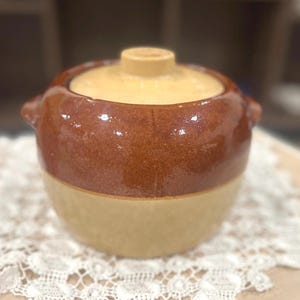 May include: A two-toned ceramic cooking pot with a lid. The upper portion is a glossy, dark brown, while the lower section and lid are a lighter tan color. The pot has small handles on either side and sits on a white lace doily.