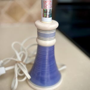 May include: A small, ceramic lamp with a blue, white, and cream color scheme. The lamp has a cylindrical base with horizontal ridges and a bulb socket. The lamp's cord is white. The text on the bulb reads "CAUTION THE RISK 15 WATT OR TYPE B".