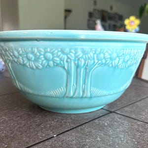 May include: A turquoise ceramic mixing bowl with a detailed embossed floral and tree design. The bowl has a wide rim and a rounded shape, suitable for kitchen use. The embossed pattern adds a decorative element to the functional item.